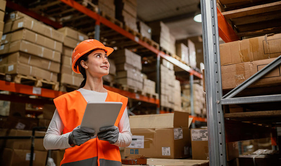 Woman in hardhat holding tablet and looking at merchandise on warehouse shelves